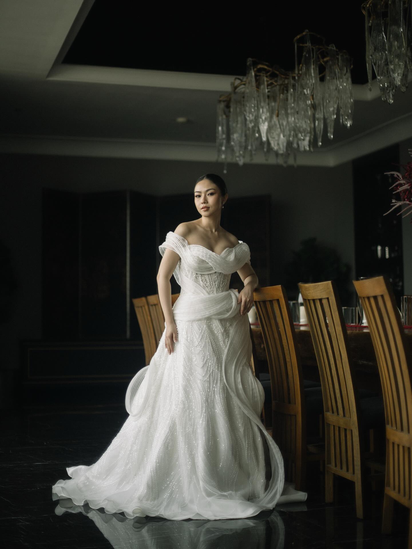 Bride in a sculptural off-shoulder wedding gown in a dark dining room by CELRA