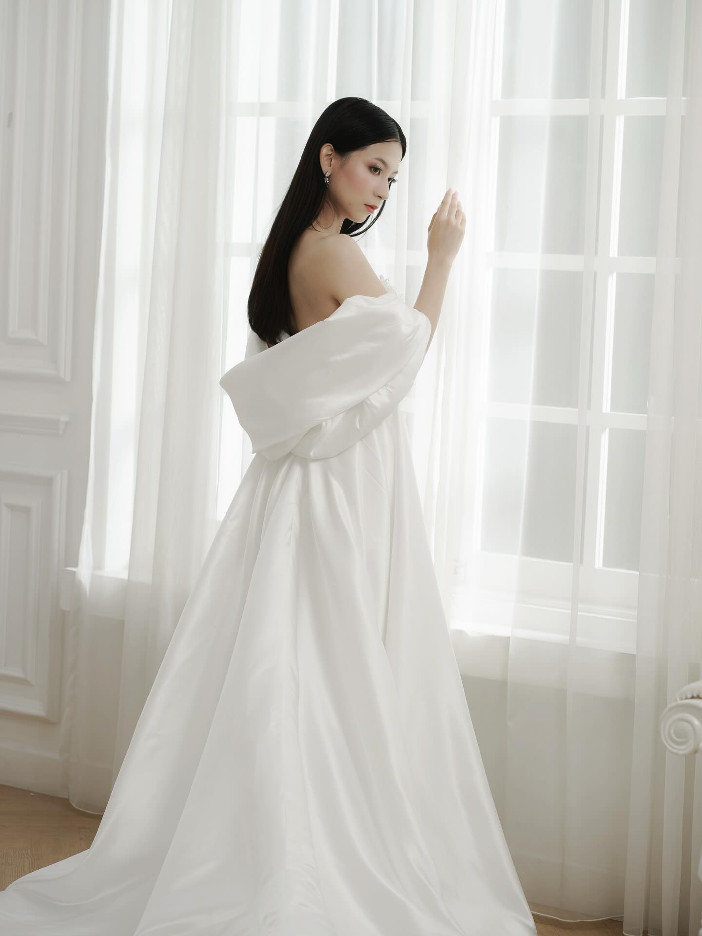 Bride in an off-shoulder lace wedding gown by a bright window by CELRA