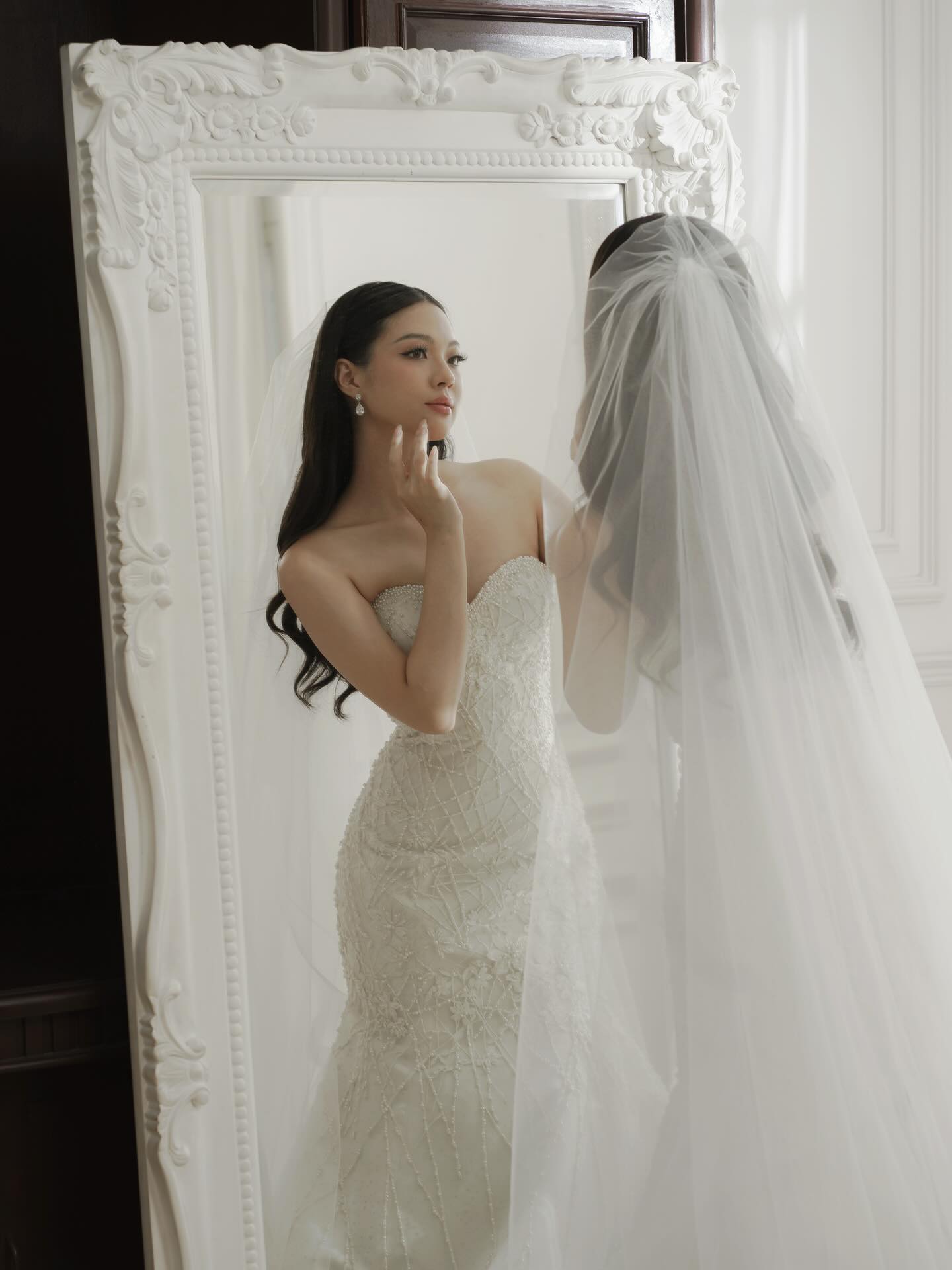 Bride in a strapless lace gown adjusting her veil before a mirror by CELRA